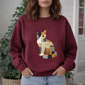 May include: Burgundy sweatshirt featuring a patchwork cat design. The cat illustration is composed of various fabric squares in shades of beige, brown, black, and orange. The sweatshirt has a crew neck and long sleeves.