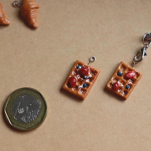 Puede incluir: Dos pequeñas gofres cuadradas marrones con coberturas rojas y azules. Las gofres están unidas a cadenas plateadas y cuelgan de una superficie marrón. Una moneda plateada está en la esquina inferior izquierda de la imagen.