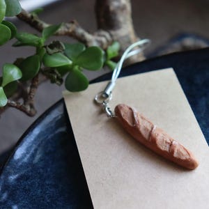Puede incluir: Un amuleto de baguette marrón unido a una cadena plateada. El amuleto está sobre un fondo de papel marrón.