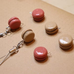 May include: Five macaron keychains, two pink and three beige, with silver metal rings and chains. The keychains are on a brown surface.
