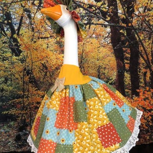 May include: A white goose figurine wearing a patchwork dress and a matching bonnet. The dress features a yellow collar with a small burlap bow, a skirt with a multi-colored floral and polka dot pattern, and a white lace trim along the bottom edge.