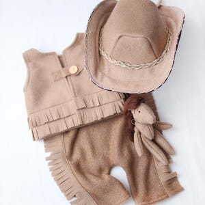 Può includere: Un completo da cowboy beige per neonati, composto da un gilet con frange, pantaloni con frange e un cappello da cowboy con bordatura in corda. È incluso anche un piccolo cavallo di peluche.