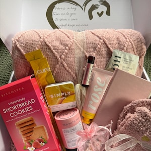 May include: A gift box filled with self-care items. Includes a pink pillow, strawberry shortbread cookies, lip balm, hand cream, tea, mints, a notebook, and a pair of socks. The box has a heart design with the words "Here's a hug from me to you."