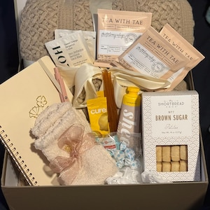 May include: A gift box containing self-care products. It features a beige knitted blanket, tea sachets, a notebook, a pen, socks with a pink bow, lotion, and a box of brown sugar shortbread biscuits.