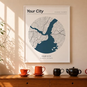 Puede incluir: Una lámina de arte enmarcada con un diseño de mapa circular que muestra una masa de agua azul oscuro y tierra gris claro. El texto 'Your City' y las coordenadas '03.0600°N / 03.0300°E' están impresos en negro en la parte superior e inferior.