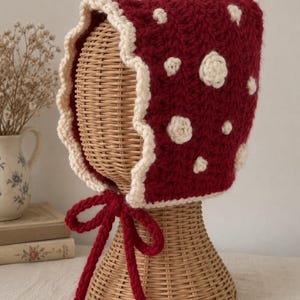 Peut inclure: Un bonnet en crochet rouge foncé avec un bord festonné de couleur crème et de petites taches circulaires crème réparties sur la surface. Le bonnet comprend de longs liens en crochet rouge foncé assortis qui pendent sous le menton.