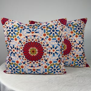 May include: Two square throw pillows featuring a cream-colored base with a vibrant, intricate geometric pattern. The design includes large red circular medallions surrounded by blue, green, and orange star-shaped motifs and complex tile-like shapes.