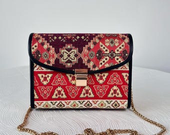 May include: A rectangular structured handbag featuring a woven tapestry fabric with a geometric pattern in shades of red, burgundy, and cream. The bag has black leather-like trim, a gold-tone metal clasp closure, and a gold-tone metal chain strap.
