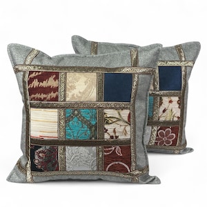 Puede incluir: Dos cojines cuadrados decorativos con un diseño de patchwork. Los cojines tienen un borde gris con ribete dorado y un panel central de varios cuadrados de tela en tonos rojos, azules, beige y turquesa. Los cojines están sobre un fondo blanco.