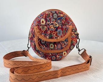May include: A round crossbody bag featuring a vibrant red fabric with a multi-colored floral and paisley pattern. The bag includes a front flap pocket with tan trim and a matching tan adjustable shoulder strap decorated with a black zigzag stitch.