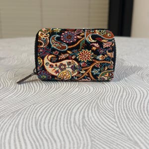 May include: A rectangular zippered wallet featuring a black base fabric with a multi-colored woven paisley and floral pattern in shades of red, blue, yellow, and green. The wallet has a black zipper closure and a small, dark-colored zipper pull tab.