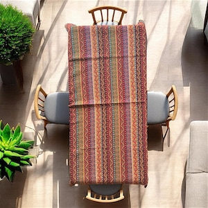 Pode incluir: Um corredor de mesa retangular com um padrão listrado colorido. O corredor apresenta um design geométrico repetitivo em tons de vermelho, laranja, amarelo, verde, azul e roxo. O corredor está colocado sobre uma mesa de madeira com duas cadeiras.