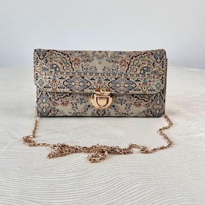 May include: A rectangular clutch bag featuring a beige fabric with a multi-colored intricate floral and geometric pattern. The bag has a gold-tone metal push-lock closure on the front flap and a matching gold-tone metal chain shoulder strap.