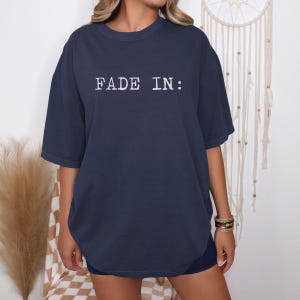 May include: Navy blue t-shirt with the text "FADE IN:" in white, centered on the chest. The shirt has short sleeves and a relaxed fit. The model is wearing the shirt with dark shorts.