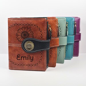 May include: A row of four bi-fold wallets in brown, light green, teal, and purple. The front wallet is brown with a sunflower engraving, the name Emily, and a black snap closure featuring a metallic flower button. Each wallet has perforated details.
