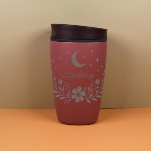 Lifestyle image of a custom name tumbler resting on a desk, featuring a floral celestial theme with moon and star elements, ideal for coffee lovers who enjoy aesthetic and personalized drinkware.