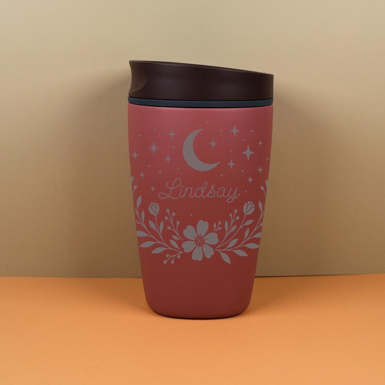 Lifestyle image of a custom name tumbler resting on a desk, featuring a floral celestial theme with moon and star elements, ideal for coffee lovers who enjoy aesthetic and personalized drinkware.