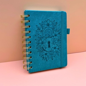May include: Teal spiral-bound notebook with a floral and keyhole design embossed on the cover. The notebook has a gold spiral binding and a teal ribbon bookmark. The background is a soft pink and peach.