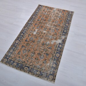 May include: Rectangular rug with a distressed, vintage appearance. The rug features a warm, orange-brown central field with floral patterns in blue and cream. A dark blue border frames the design. The rug is placed on a light-colored surface.