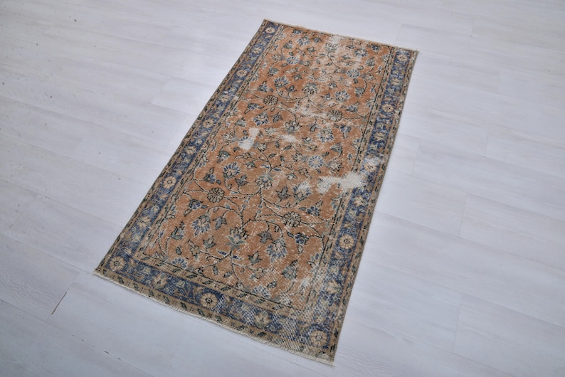 May include: Rectangular rug with a distressed, vintage appearance. The rug features a warm, orange-brown central field with floral patterns in blue and cream. A dark blue border frames the design. The rug is placed on a light-colored surface.