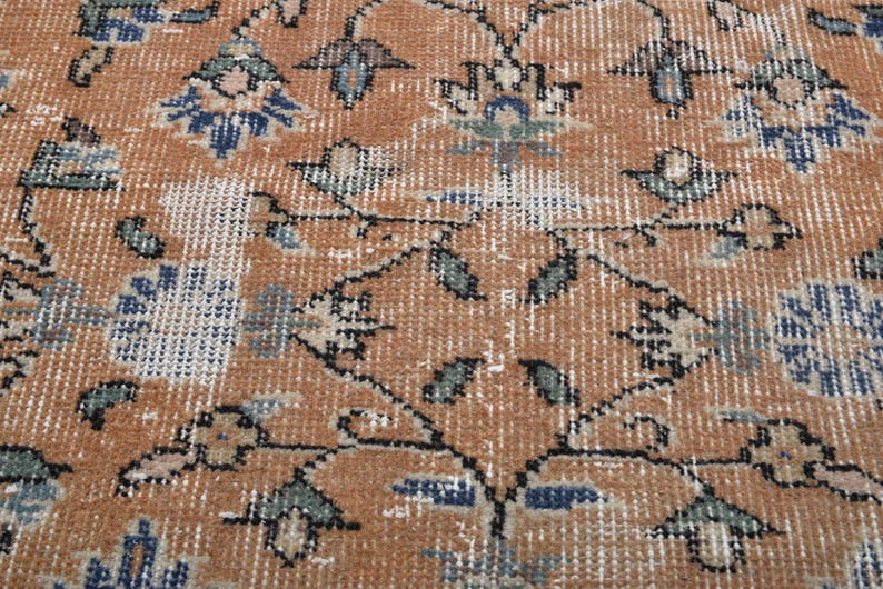 May include: Close-up of a distressed, vintage rug with a warm, earthy color palette. The rug features a repeating floral pattern in shades of rust, blue, and cream, with intricate details and a worn texture. The design has a classic, traditional feel.
