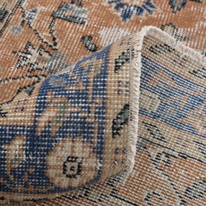 May include: Close-up of a section of a vintage rug with a folded corner. The rug features a distressed design with a color palette of tan, navy blue, and touches of green. The pattern includes floral and geometric elements, creating a classic, aged aesthetic.