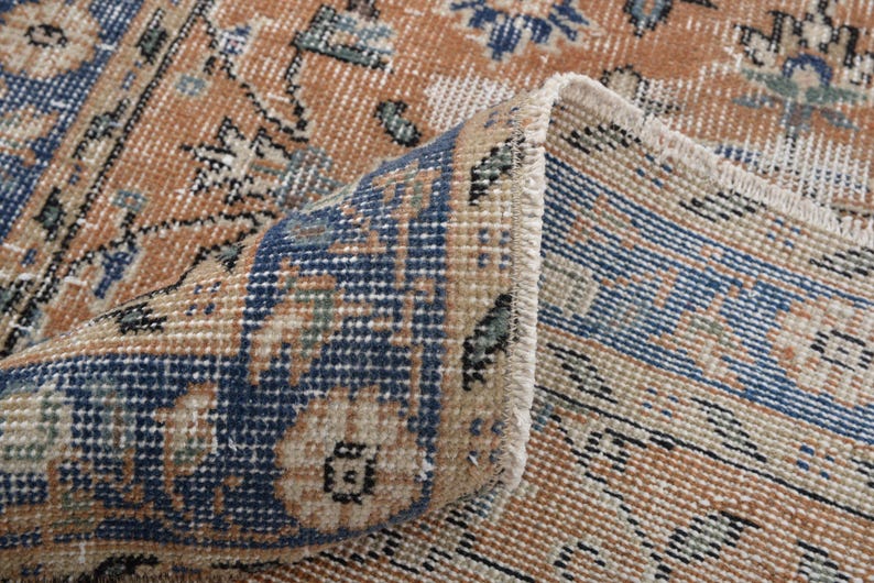 May include: Close-up of a section of a vintage rug with a folded corner. The rug features a distressed design with a color palette of tan, navy blue, and touches of green. The pattern includes floral and geometric elements, creating a classic, aged aesthetic.