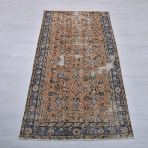 May include: Rectangular vintage rug with a distressed design. The rug features a central field of tan with floral patterns in blue and cream. A dark blue border frames the design. The rug is placed on a light-colored wooden floor.