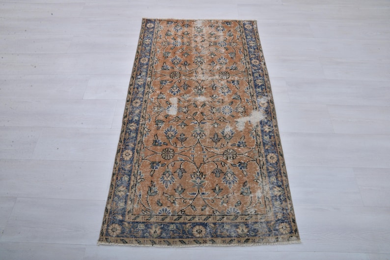 May include: Rectangular vintage rug with a distressed design. The rug features a central field of tan with floral patterns in blue and cream. A dark blue border frames the design. The rug is placed on a light-colored wooden floor.