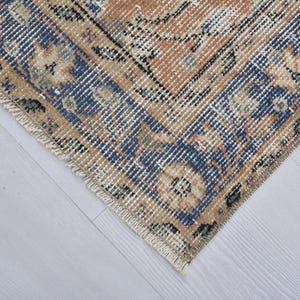 May include: Close-up of a section of a vintage rug with a distressed, faded appearance. The rug features a central design in shades of tan and blue, with floral and geometric patterns. The edges have a fringed border.