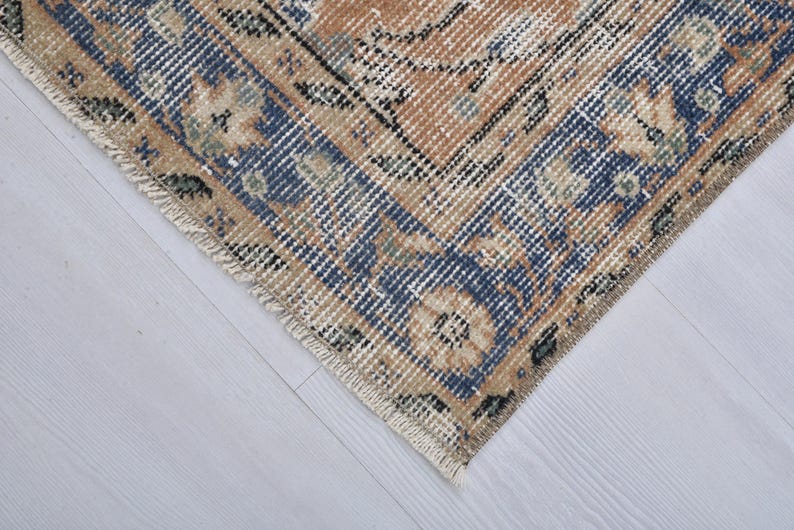 May include: Close-up of a section of a vintage rug with a distressed, faded appearance. The rug features a central design in shades of tan and blue, with floral and geometric patterns. The edges have a fringed border.