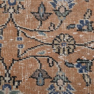 May include: Close-up of a distressed, vintage rug with an intricate floral pattern. The rug features a warm, orange-brown background with detailed designs in blue, black, and white. The rug's texture and color palette suggest a traditional, antique style.