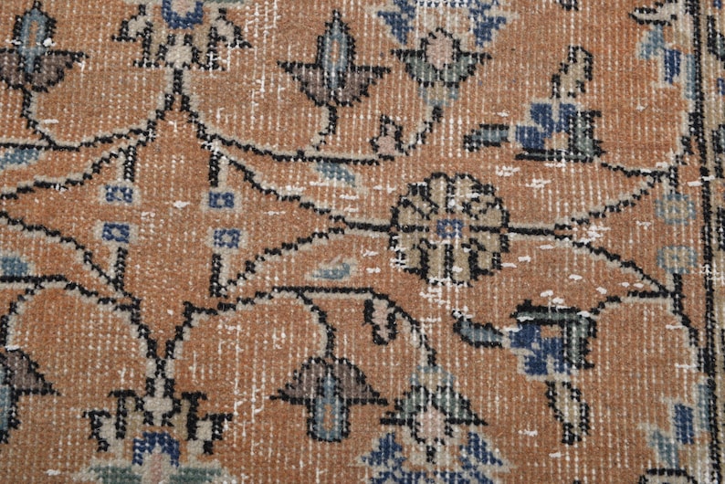 May include: Close-up of a distressed, vintage rug with an intricate floral pattern. The rug features a warm, orange-brown background with detailed designs in blue, black, and white. The rug's texture and color palette suggest a traditional, antique style.