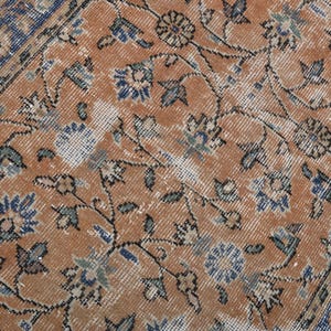 May include: Close-up of a distressed, vintage rug with an intricate floral pattern. The rug features a warm, orange-brown central field with blue and green floral motifs and a blue border. The design has a faded, antique appearance.
