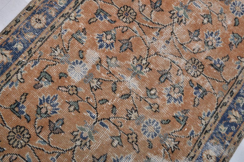 May include: Close-up of a distressed, vintage rug with an intricate floral pattern. The rug features a warm, orange-brown central field with blue and green floral motifs and a blue border. The design has a faded, antique appearance.