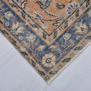 May include: Close-up of a section of a vintage rug with a distressed appearance. The rug features a central design in shades of peach and beige, framed by a dark blue border with floral patterns. The rug's edges show signs of wear.