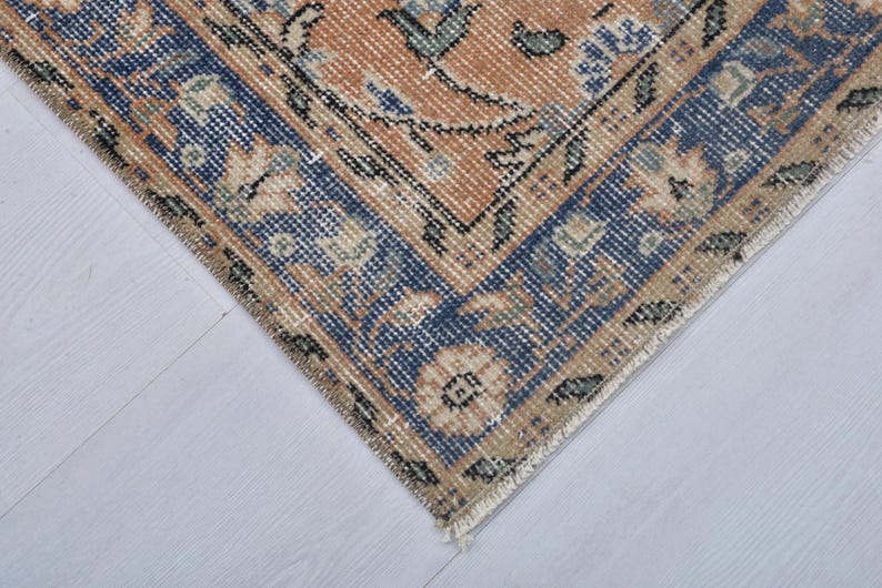 May include: Close-up of a section of a vintage rug with a distressed appearance. The rug features a central design in shades of peach and beige, framed by a dark blue border with floral patterns. The rug's edges show signs of wear.