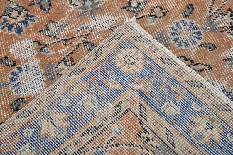 May include: Close-up of a section of a vintage rug with a distressed, faded appearance. The rug features a warm, earthy color palette with shades of brown, beige, and blue. The design includes floral and geometric patterns, with a visible fringe.
