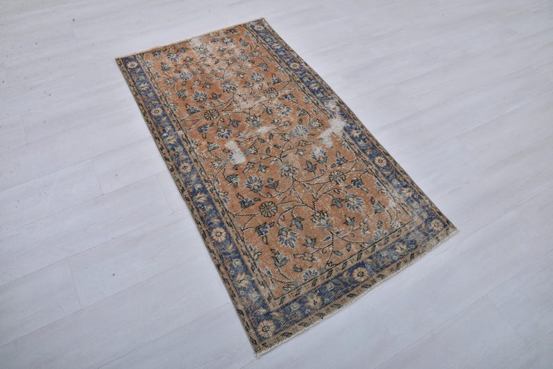 May include: Rectangular rug with a distressed, vintage look. The rug features a central field of warm orange with floral patterns in blue and cream. A dark blue border frames the design, adding contrast. Ideal for living rooms or bedrooms.