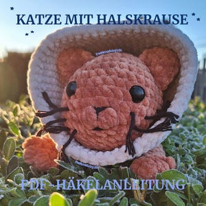 May include: A handmade, plush cat toy in shades of peach and cream, wearing a white and cream hat. The cat has black eyes and whiskers. Text on the image reads "KATZE MIT HALSKRAUSE" and "PDF HÄKELANLEITUNG".