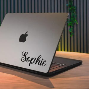 Op de afbeelding: Een zilveren laptop met het Apple-logo en de naam "Sophie" in zwart script. De laptop staat open op een houten oppervlak, met een vage achtergrond van verticale lijnen en groen.