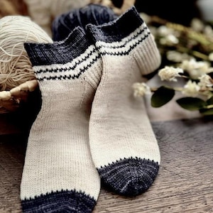 Puede incluir: Un par de calcetines de punto a la altura del tobillo con cuerpo de color crema y punteras y puños de color gris carbón oscuro. Los puños presentan dos finas rayas horizontales en color crema que contrastan con el tejido gris oscuro.