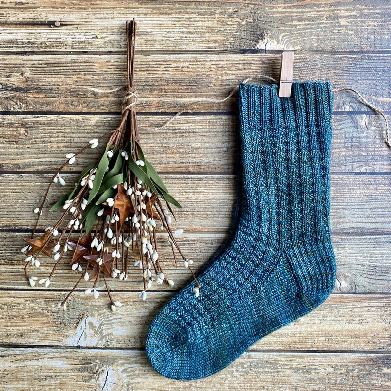 May include: A teal blue knitted sock with a ribbed texture. The sock is hanging from a wooden clothespin on a wood background.