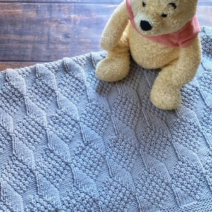 May include: A gray knitted baby blanket with a diamond pattern. A plush Winnie the Pooh stuffed animal is sitting on the blanket.