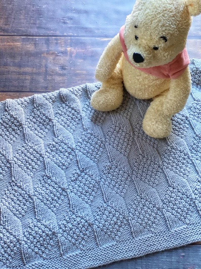 May include: A gray knitted baby blanket with a diamond pattern. A plush Winnie the Pooh stuffed animal is sitting on the blanket.