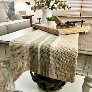 May include: A beige and green striped knitted table runner on a wooden coffee table. The table runner is made of a soft, textured yarn and has a subtle sheen. The table runner is a great way to add a touch of warmth and style to your home decor.