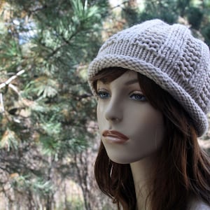 May include: A close-up of a woman wearing a beige knitted beanie hat. The hat has a simple, textured design and covers her ears. Her hair is long and brown, and she is looking off to the side.