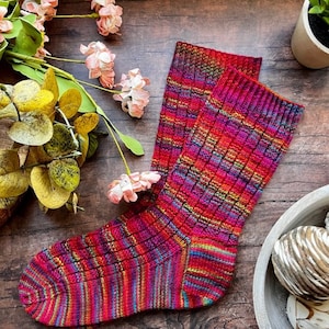 May include: A pair of colorful striped knitted socks with a textured pattern. The socks are made with a variety of shades of red, pink, orange, and purple yarn.