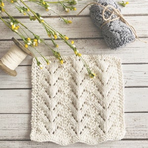 May include: A white knitted dishcloth with a lacy pattern. The dishcloth is lying on a white wooden surface with a spool of yarn and a ball of yarn in the background.
