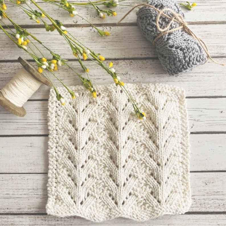May include: A white knitted dishcloth with a lacy pattern. The dishcloth is lying on a white wooden surface with a spool of yarn and a ball of yarn in the background.